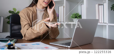Young Asian business woman sits on the phone in an online business meeting using a laptop in a modern home office decorated with shady green plants. Young Asian business woman sits on the phone in an online business meeting using a laptop in a modern home office decorated with shady green plants. 122255316