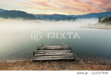 Misty morning view of a rustic wooden pier on a calm lake with a mountainous backdrop 122257930