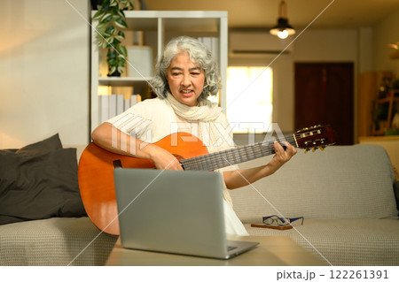 Happy senior woman practicing guitar in living room and following an online lesson on laptop Happy senior woman practicing guitar in living room and following an online lesson on laptop 122261391