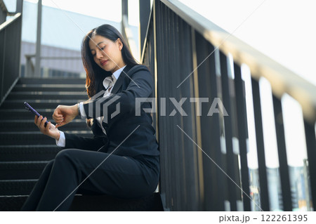 Businesswoman in a suit checking the time on her watch while using phone on the stairs in the city Businesswoman in a suit checking the time on her watch while using phone on the stairs in the city 122261395