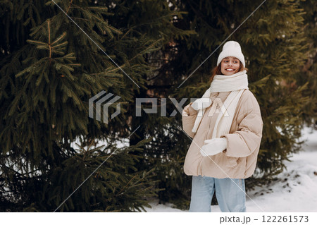 Happy smiling young woman portrait dressed coat scarf hat and mittens enjoys winter weather at winter park with pines 122261573
