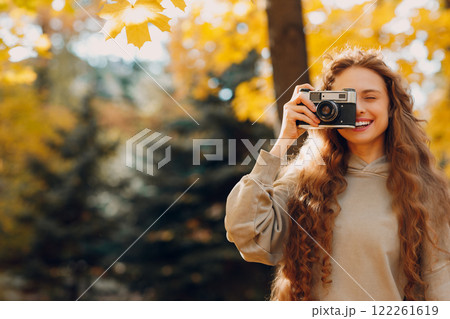 Smiling young woman with the photo camera shooting the forest trees with the yellow leaves at sunset 122261619