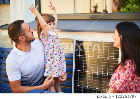 Happy family spending time together with solar panel. Father, holding young girl, sits on bench with mother. Scene highlights importance of educating next generation about renewable energy. 122263797
