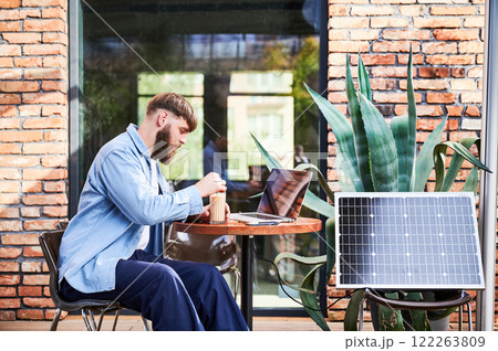 Man works on laptop at outdoor round table, with glass of iced coffee. Solar panel nearby, emphasizing sustainable, modern eco-friendly workspace that combines technology and sustainability. 122263809