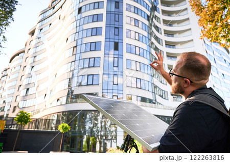 Man holding photovoltaic solar panel in front of new building, showing Ok sign. Concept of integration of sustainable renewable energy sources into modern architecture. Man holding photovoltaic solar panel in front of new building, showing Ok sign. Concept of integration of sustainable renewable energy sources into modern architecture. 122263816