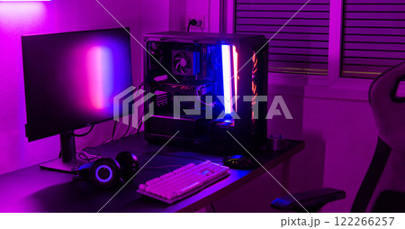 A computer setup with a purple light dark background, games transmission modern equipment, modern gaming PC on desk with keyboard mouse monitor, gamer work space concept 122266257