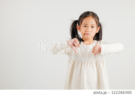 Portrait disheartened Asian young kid girl displaying a thumbs down gesture, symbolizing dislike. Studio shot isolated on white with copy space, kindergarten child portraying negative and sad look 122266300