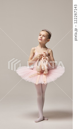 Young ballerina, wearing pink tutu and tiara, posing elegantly holding pointe shoes with confident gaze against white studio background. 122267143