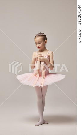 Little ballerina, wearing pink tutu and tiara, standing calmly, with closed eyes, holding pointe shoes in hands against white studio background. 122267144
