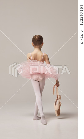 Back view of little ballet dancer in pink tutu holds ballet pointe, walking away gracefully, embodying reflective moment against beige studio background. 122267168