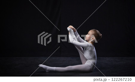 Young ballerina holding one leg extended high with arms, displaying balance and flexibility, dressed in white suit, against black studio background. 122267210
