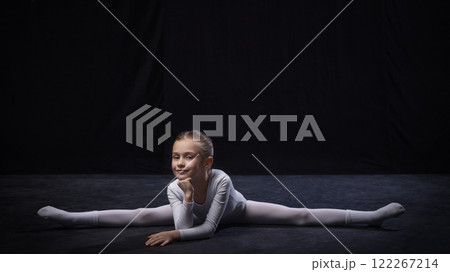 Kid, ballerina smiling while holding forward split, hands on chin, dressed in white, expressing flexibility and poise, against black studio background. 122267214