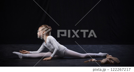 Young ballerina stretching in forward split, focused expression, dressed in white ballet attire, against black studio background. Young ballerina stretching in forward split, focused expression, dressed in white ballet attire, against black studio background. 122267217