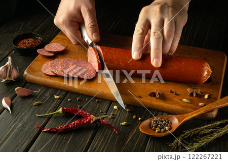 In a warm, inviting kitchen, hands expertly cut through a log of sausage on a wooden cutting board. Surrounding spices and garlic create a vibrant culinary atmosphere, perfect for food lovers 122267221