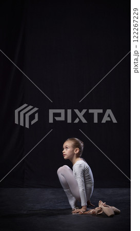 Young ballerina squatting on floor, gazing upward thoughtfully, wearing white costume with pointe shoes placed beside, against black background. Young ballerina squatting on floor, gazing upward thoughtfully, wearing white costume with pointe shoes placed beside, against black background. 122267229