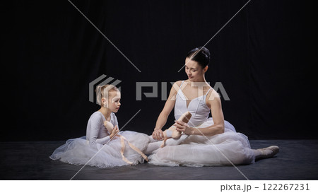 Adult ballerina sitting cross-legged helping child with ballet shoes, both in white costumes, sharing moment, against black studio background. 122267231