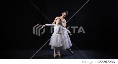 Adult ballerina guiding child in ballet posture, gently supporting arms, both dressed in white tutus, against black studio background. 122267233