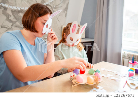 Side view of little Caucasian girl and her mother with homemade paper bunny masks are having fun playing in the kitchen.Family sits at table and makes festive Easter decorations. Easter Celebration 122267673