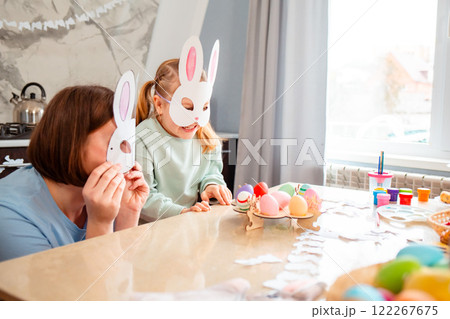 Low angle view of little Caucasian girl and her mom with homemade paper bunny masks are having fun playing in the kitchen. A family sits at a table and makes festive Easter decorations 122267675