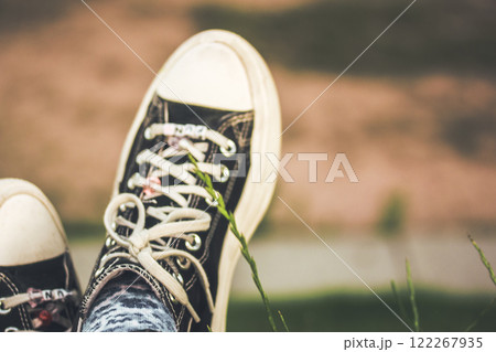 A young girl in black and white sneakers relaxing sitting on a green lawn in summer. Rest in nature. Green grass in a garden, park. Girlish footwear. 122267935