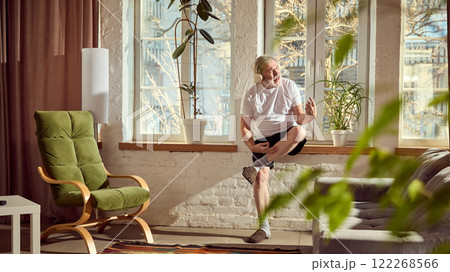 Elderly man in home wear listening to music in headphones , sitting on windowsill and playing imaginative guitar in cozy, warmly lit living room at home Elderly man in home wear listening to music in headphones , sitting on windowsill and playing imaginative guitar in cozy, warmly lit living room at home 122268566