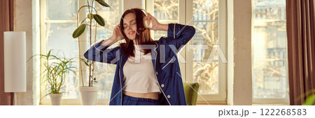 Young relaxed smiling woman in blue silk pajamas, listening to music in headphones with positive expression, relaxing at home in warmly lit living room Young relaxed smiling woman in blue silk pajamas, listening to music in headphones with positive expression, relaxing at home in warmly lit living room 122268583
