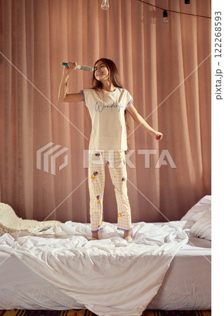 Home concert. Happy, smiling teen girl in pajamas standing on bed and singing in hairbrush microphone. Cozy bedroom and home atmosphere Home concert. Happy, smiling teen girl in pajamas standing on bed and singing in hairbrush microphone. Cozy bedroom and home atmosphere 122268593