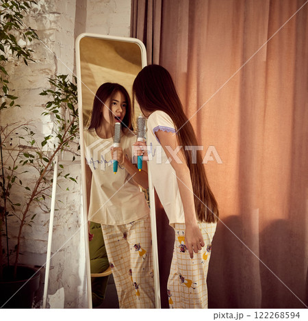 Teen girl in cute pajamas looking in mirror and singing with hairbrush, at home, in cozy room with white brick walls and plants Teen girl in cute pajamas looking in mirror and singing with hairbrush, at home, in cozy room with white brick walls and plants 122268594