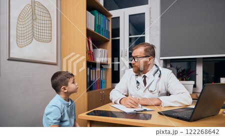 Little boy, child in blue t-shirt visiting doctor in medical office, sitting and talking to bearded man in lab coat listening with attention and care Little boy, child in blue t-shirt visiting doctor in medical office, sitting and talking to bearded man in lab coat listening with attention and care 122268642