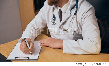 Cropped image of bearded man , doctor in white coat and stethoscope sitting at table and prescribing treatment for patient, taking notes 122268643