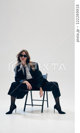 Businesswoman in sunglasses, sitting in confident pose on chair, in stylish formal wear with black costume, white shirt and a tie against studio background 122269918