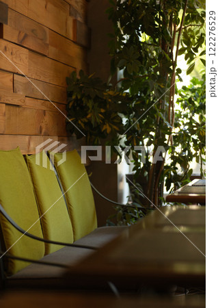 Cozy interior of a cafe with a wooden wall, green cushioned chairs, and lush indoor plants creating a relaxing and inviting atmosphere. Cozy interior of a cafe with a wooden wall, green cushioned chairs, and lush indoor plants creating a relaxing and inviting atmosphere. 122276529