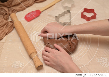 Christmas homemade baking, woman kneading brown cocoa dough with her hands for baking gingerbread, shaped cutter, silicone spatula and rolling pin on the table Christmas homemade baking, woman kneading brown cocoa dough with her hands for baking gingerbread, shaped cutter, silicone spatula and rolling pin on the table 122277099