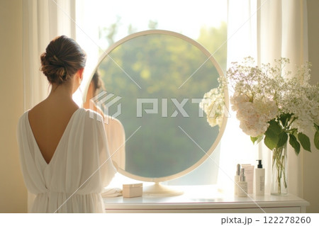 A woman in her late 20s, with her hair styled in a low bun, stands in front of a vanity mirror 122278260