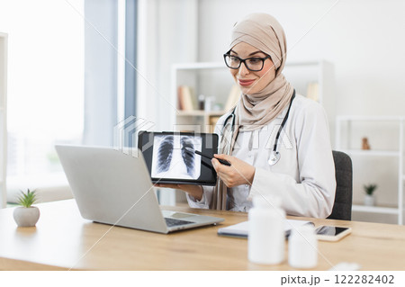 Muslim female doctor in early 30s conducts virtual consultation showing lung x-ray to patient. Professional and focused medical setup with laptop and stethoscope in modern office environment. 122282402