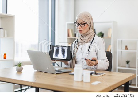 Young Muslim female doctor having video call with patient showing x-ray of diseased lung. Professional working in modern office setting, focused on telemedicine and patient care. Young Muslim female doctor having video call with patient showing x-ray of diseased lung. Professional working in modern office setting, focused on telemedicine and patient care. 122282405