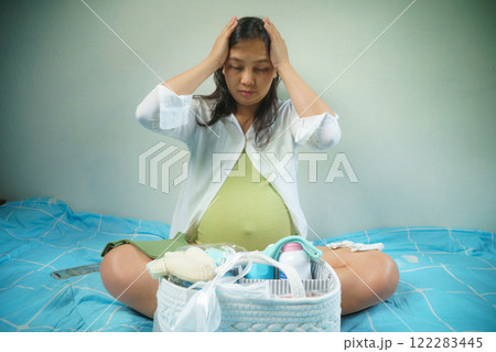 pregnant woman sits on bed, holding her head in stress, surrounded by baby essentials in basket, symbolizing preparation and emotional challenges during pregnancy pregnant woman sits on bed, holding her head in stress, surrounded by baby essentials in basket, symbolizing preparation and emotional challenges during pregnancy 122283445