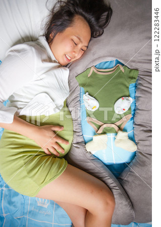 pregnant woman lying on bed, smiling joyfully while holding her belly, with green baby onesie and knitted baby booties placed beside her, symbolizing anticipation and love pregnant woman lying on bed, smiling joyfully while holding her belly, with green baby onesie and knitted baby booties placed beside her, symbolizing anticipation and love 122283446