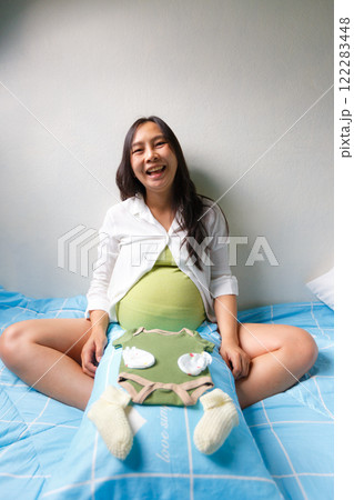 pregnant woman sitting on bed with joyful expression, holding baby clothes and accessories on her belly, symbolizing excitement and preparation for motherhood 122283448