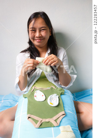 smiling woman sits on bed holding knitted baby sock, with baby clothes and accessories placed on her lap, creating warm and joyful atmosphere 122283457