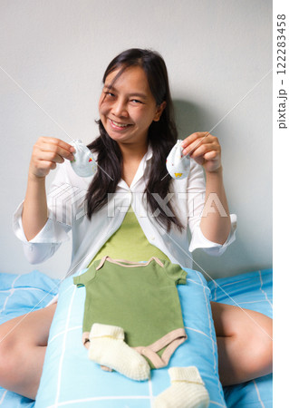 smiling pregnant woman sits on bed holding baby socks, with green baby onesie and yellow socks placed on pillow in front of her, expressing joy and anticipation smiling pregnant woman sits on bed holding baby socks, with green baby onesie and yellow socks placed on pillow in front of her, expressing joy and anticipation 122283458