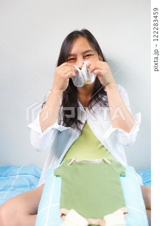 pregnant woman sitting on bed, smiling joyfully while holding baby socks, with green baby onesie placed on her lap, expressing excitement and anticipation for her baby 122283459