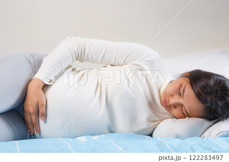 woman lying on bed, holding her belly, showing signs of discomfort. She is wearing long sleeve top and leggings, with calm expression woman lying on bed, holding her belly, showing signs of discomfort. She is wearing long sleeve top and leggings, with calm expression 122283497