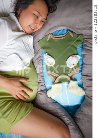 pregnant woman lies on bed, smiling while looking at baby clothes, including green onesie, mittens, and socks, symbolizing anticipation and joy for upcoming baby pregnant woman lies on bed, smiling while looking at baby clothes, including green onesie, mittens, and socks, symbolizing anticipation and joy for upcoming baby 122283518