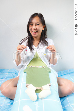 smiling woman sits on bed, holding baby socks and onesie, expressing joy and excitement while preparing baby clothes. scene conveys warmth and anticipation smiling woman sits on bed, holding baby socks and onesie, expressing joy and excitement while preparing baby clothes. scene conveys warmth and anticipation 122283523