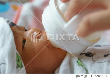 close up of newborn baby wearing white hat and blanket, being bottle fed with care and tenderness. baby is lying down, focusing on feeding bottle 122283594