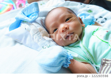 newborn baby lies on soft bed, wearing light green outfit and blue mittens, gazing curiously at camera. cozy setting is filled with warmth and gentle light 122283598