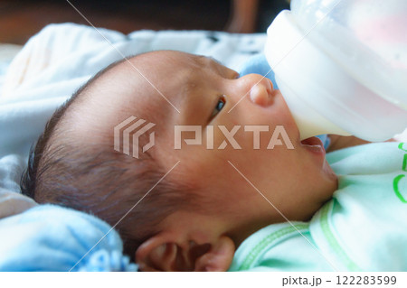 newborn baby lying down and drinking milk from bottle, wearing light green outfit, with soft and calm expression 122283599