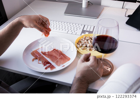 Man enjoys prosciutto, pistachios, and red wine at office desk 122293374