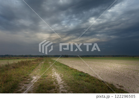 A dark stormy sky with mammatus clouds above a dirt road and fields, on a summer evening A dark stormy sky with mammatus clouds above a dirt road and fields, on a summer evening 122296505